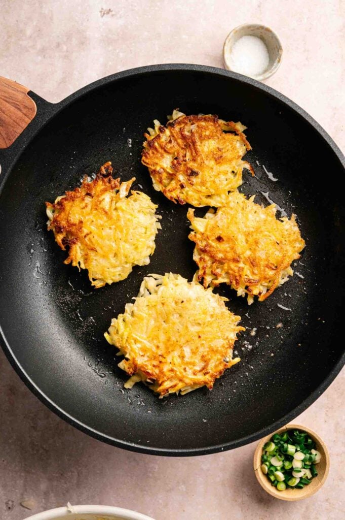 Four golden-brown potato fritters are cooking in a black frying pan. Small bowls with green onions and salt are nearby on a light-colored surface.