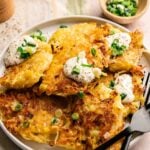 A plate of crispy potato fritters topped with sour cream and chopped green onions, with a fork and knife on the side and a small bowl of green onions in the background.