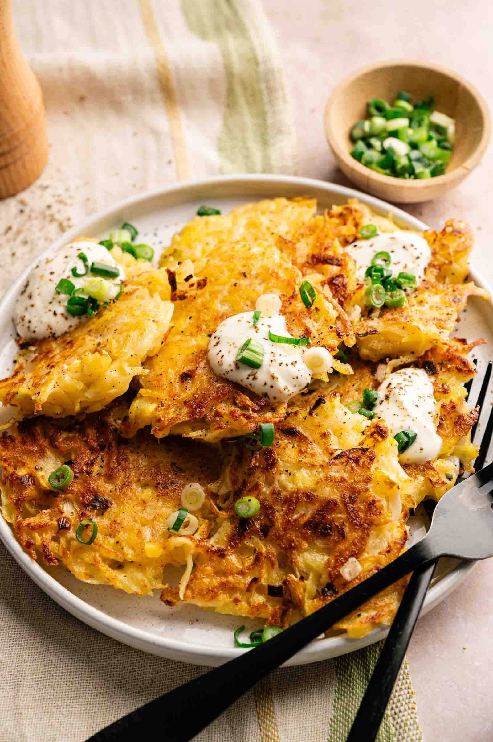 A plate of crispy potato fritters topped with sour cream and chopped green onions, with a fork and knife on the side and a small bowl of green onions in the background.