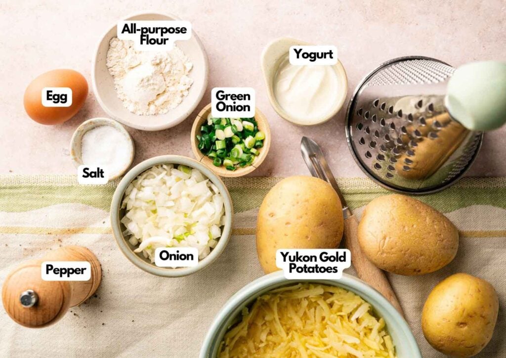 A collection of labeled ingredients for Potato Fritters on a countertop: egg, all-purpose flour, yogurt, salt, green onion, onion, pepper, and Yukon Gold potatoes with a grater.