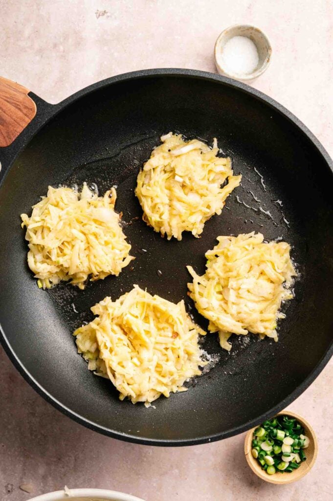 Four uncooked potato fritters in a black frying pan, with a small bowl of salt and a small bowl of chopped green onions nearby.