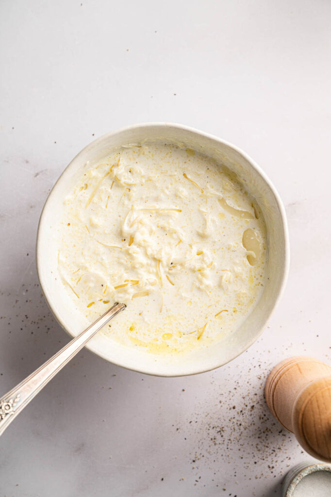 A white bowl filled with creamy sauce and shredded cheese, with a spoon inside—perfect for serving Air Fryer Potatoes Au Gratin. A wooden pepper grinder is visible nearby on the right.