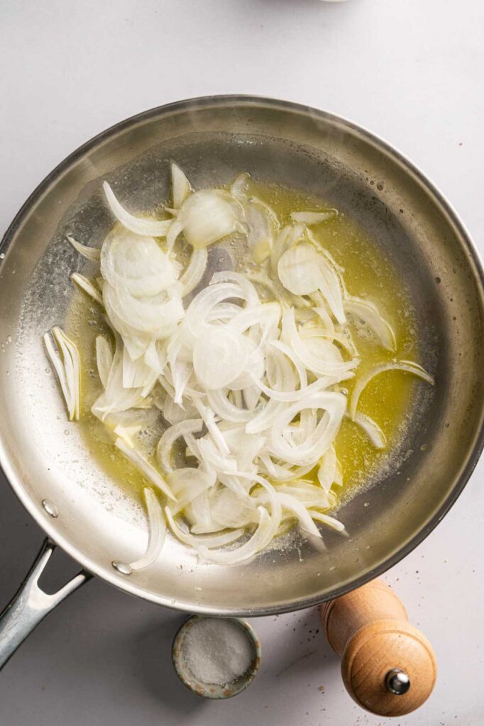Sliced onions cooking in melted butter in a stainless steel pan, with a small dish of salt and a wooden pepper grinder nearby—perfect ingredients to enhance your savory Air Fryer Potatoes Au Gratin.