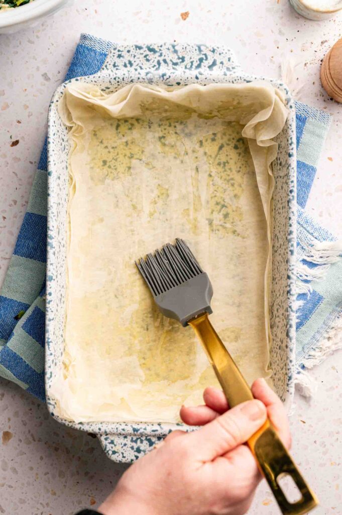 A hand brushes olive oil over layered phyllo sheets in a baking dish, prepping the base for spanakopita.