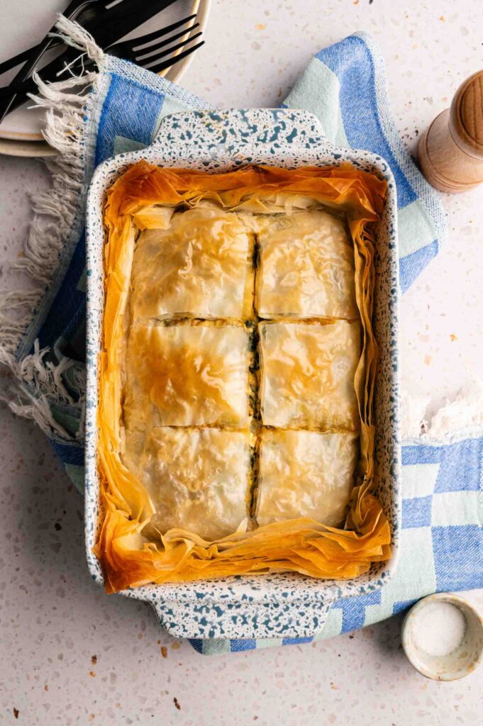 A golden tray of fresh spanakopita, cut into six pieces, sits atop a blue and white checkered towel, surrounded by plates, cutlery, and a pepper grinder for serving.