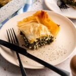 A slice of spanakopita—spinach and feta pie with flaky phyllo pastry—is served on a plate with a fork and knife, while another plate and the baking dish sit in the background.