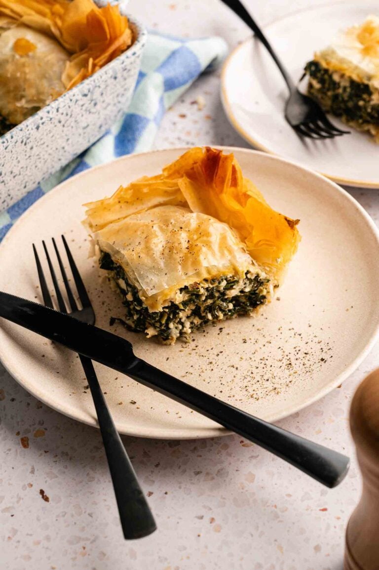 A slice of spanakopita—spinach and feta pie with flaky phyllo pastry—is served on a plate with a fork and knife, while another plate and the baking dish sit in the background.