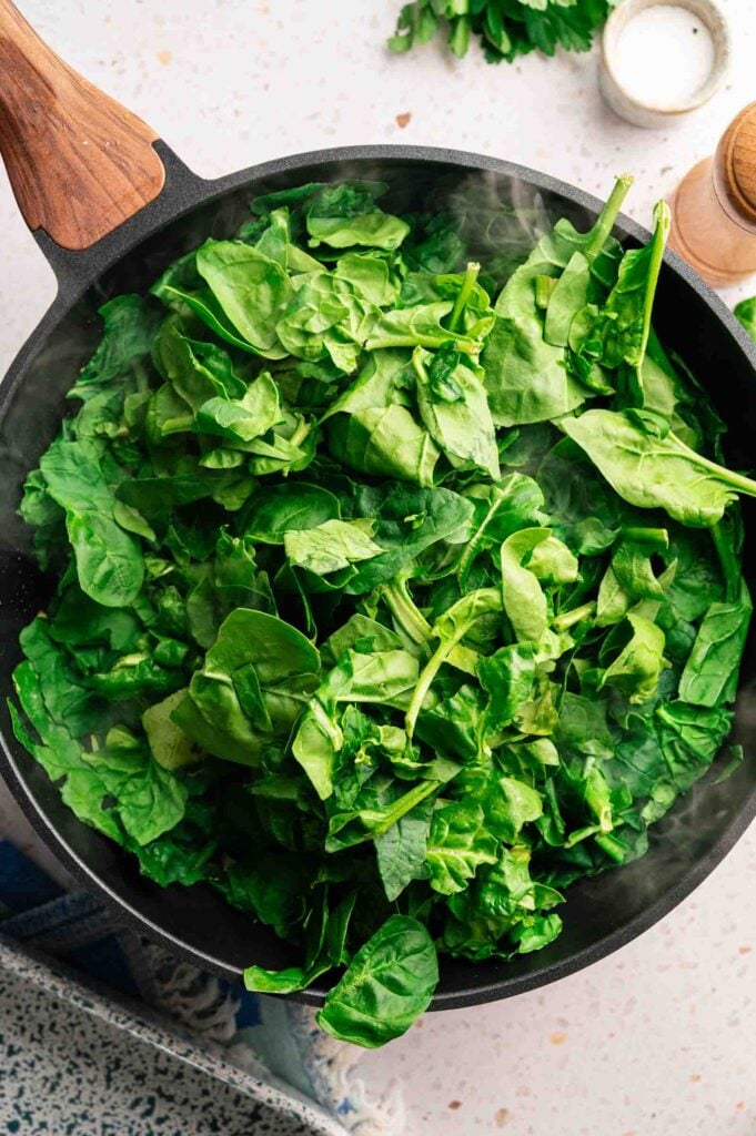 Fresh spinach leaves wilt in a skillet with steam rising, cooking down before mixing with feta.