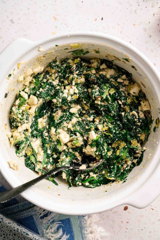 Wilted spinach is mixed with crumbled feta and fresh herbs in a bowl, forming the spanakopita filling.