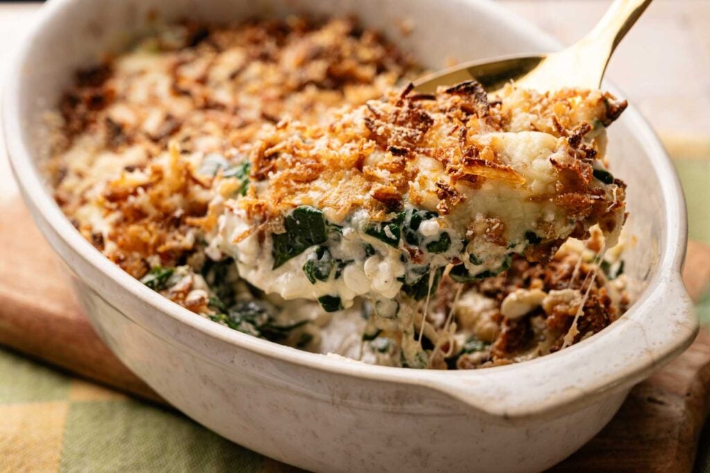 A serving spoon lifts a portion of creamy Spinach Casserole with a crispy browned topping from a white oval dish.