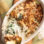 A golden, crispy Spinach Casserole with cheese is being served from an oval dish, its inviting aroma filling the air as a large spoon lifts a hearty portion.