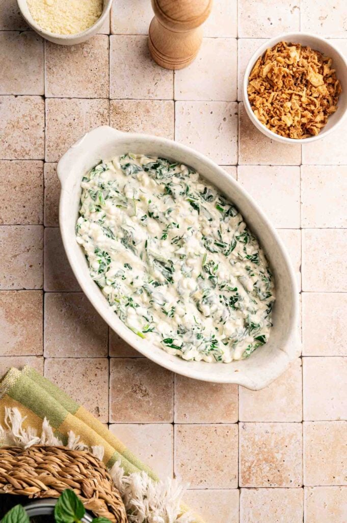 An oval dish filled with creamy spinach casserole sits on a tiled surface, with bowls of grated cheese and crispy topping nearby.