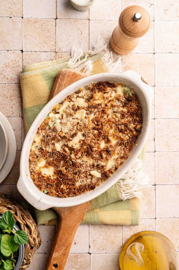 An oval dish of baked Spinach Casserole with a browned, crispy topping sits on a wooden board and checkered napkin, surrounded by a pepper grinder, olive oil, and a small plant.