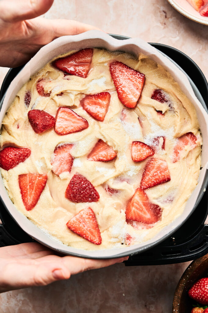 A round cake pan filled with unbaked Air Fryer Strawberry Cake batter and sliced strawberries on top, held by two hands over a light surface.