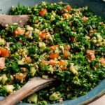 A bowl of classic tabbouleh salad featuring chopped parsley, tomatoes, cucumbers, and bulgur, all mixed with wooden utensils.
