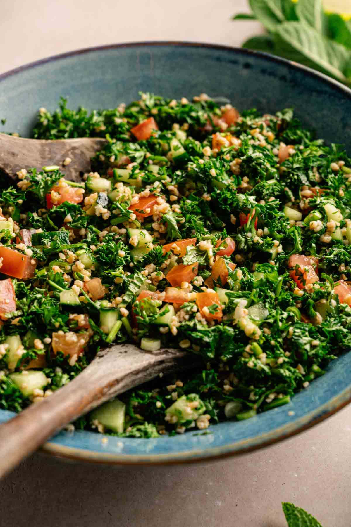 A bowl of tabbouleh salad featuring fresh chopped parsley, tomatoes, cucumber, and bulgur, all mixed together with a wooden spoon.