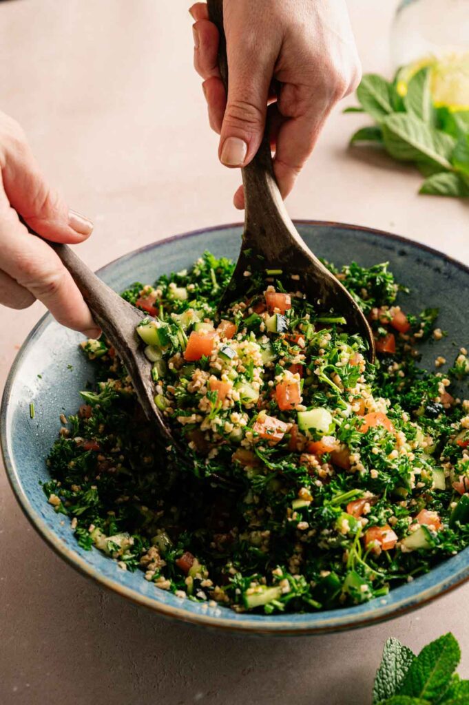Two wooden spoons toss together a bowl of fresh tabbouleh, mixing the herbs, bulgur, and vegetables.