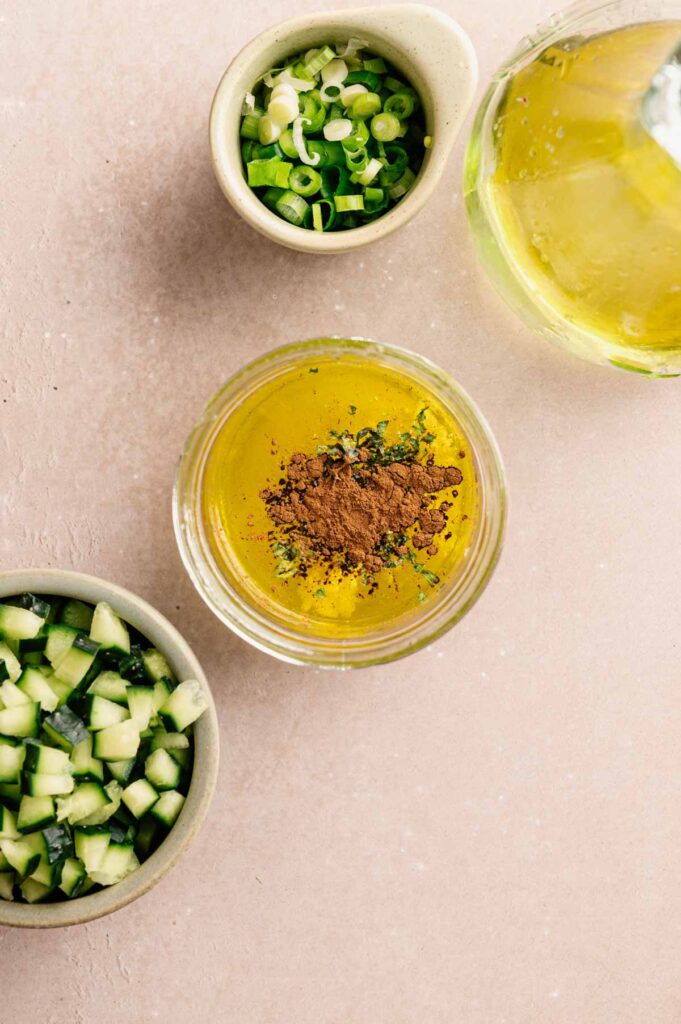 A small jar of lemon and olive oil dressing is topped with cinnamon and chopped herbs, with scallions and cucumber nearby.