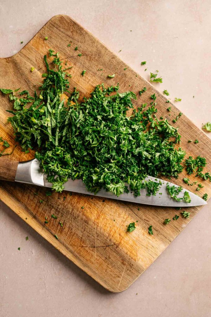 Finely chopped parsley and mint are piled on a wooden cutting board next to a chef's knife.