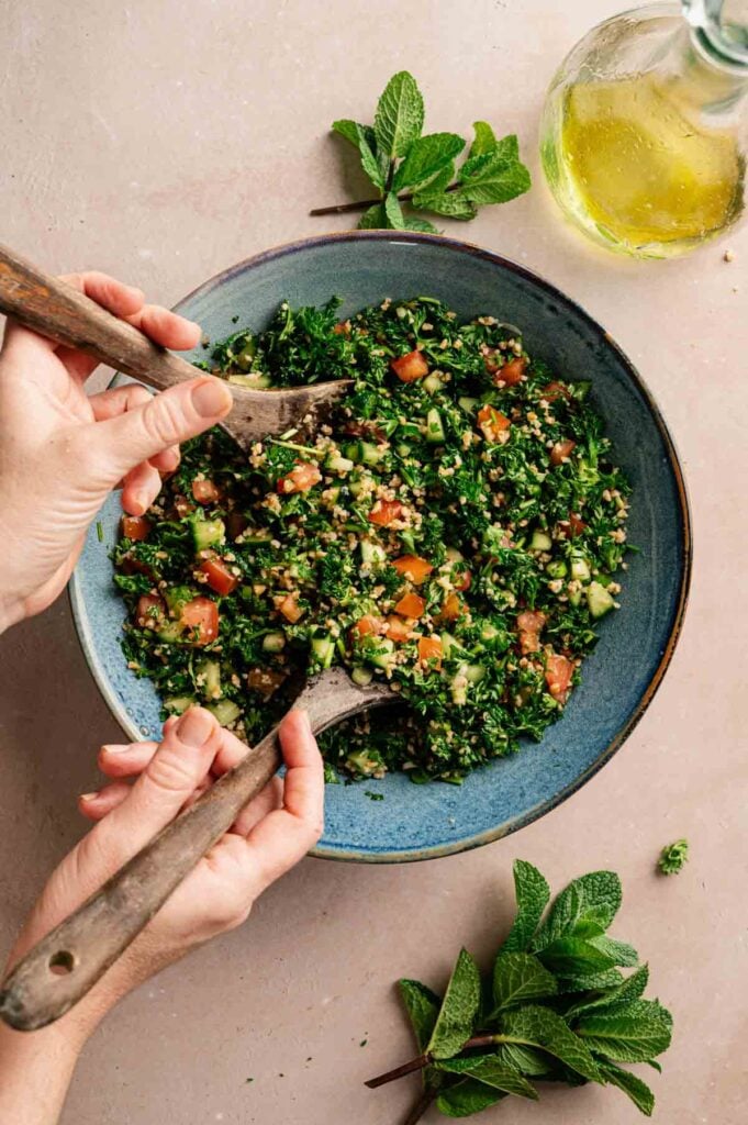 A person mixes vibrant tabbouleh salad with wooden utensils in a blue bowl; fresh mint and a bottle of olive oil sit nearby, adding to the classic tabbouleh preparation.