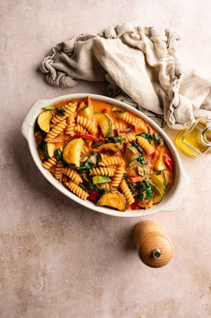 Oval dish with Air Fryer Vegetable Pasta Bake featuring short pasta, zucchini, spinach, and sauce, set on a countertop beside a beige cloth, olive oil bottle, and wooden pepper grinder.