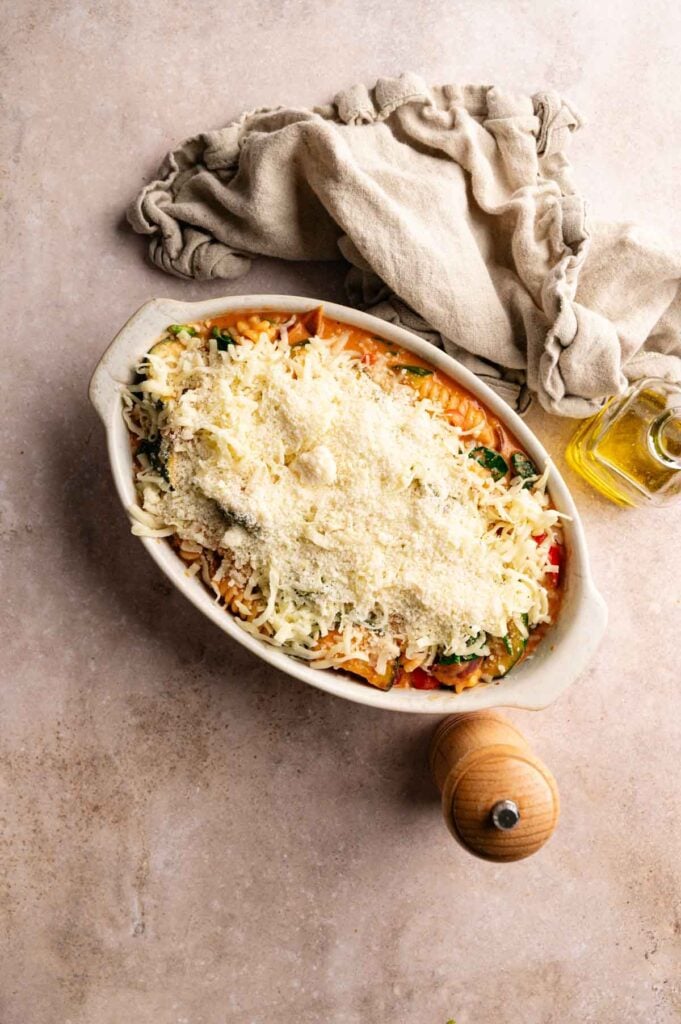 A casserole dish filled with an unbaked Air Fryer Vegetable Pasta Bake topped with shredded cheese, next to a pepper mill, olive oil bottle, and folded beige cloth.