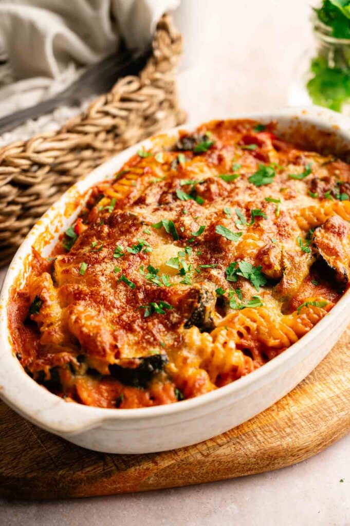 Vegetable Pasta Bake casserole topped with melted cheese and chopped parsley in a white oval dish, set on a wooden board.