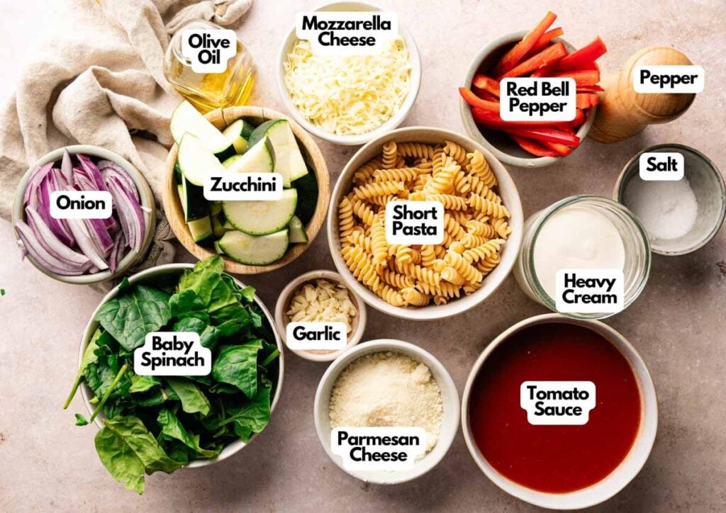 Bowls of various pasta ingredients, perfect for an Air Fryer Vegetable Pasta Bake, including short pasta, tomato sauce, mozzarella, parmesan, baby spinach, zucchini, red bell pepper, onion, garlic, heavy cream, olive oil, salt, and pepper.