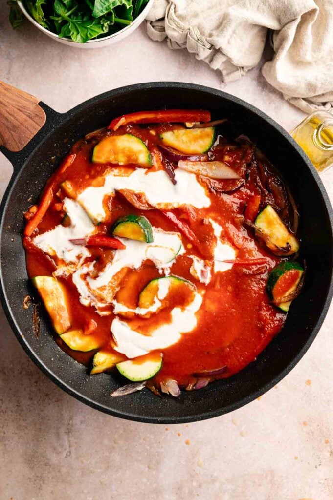 A skillet with sliced zucchini, red bell peppers, and onions in tomato sauce, topped with a swirl of cream.