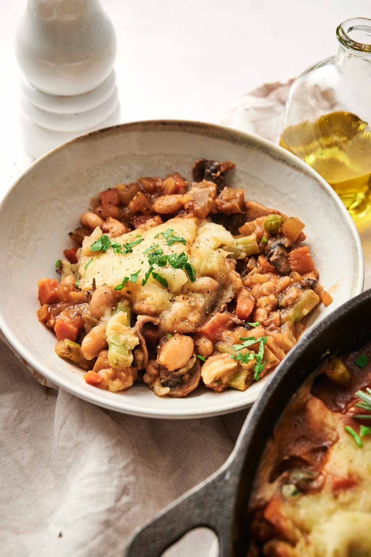 A bowl of Vegetarian Shepherd's Pie, featuring vegetable and bean stew topped with mashed potatoes and garnished with chopped parsley, sits on a white surface beside a glass bottle of oil.