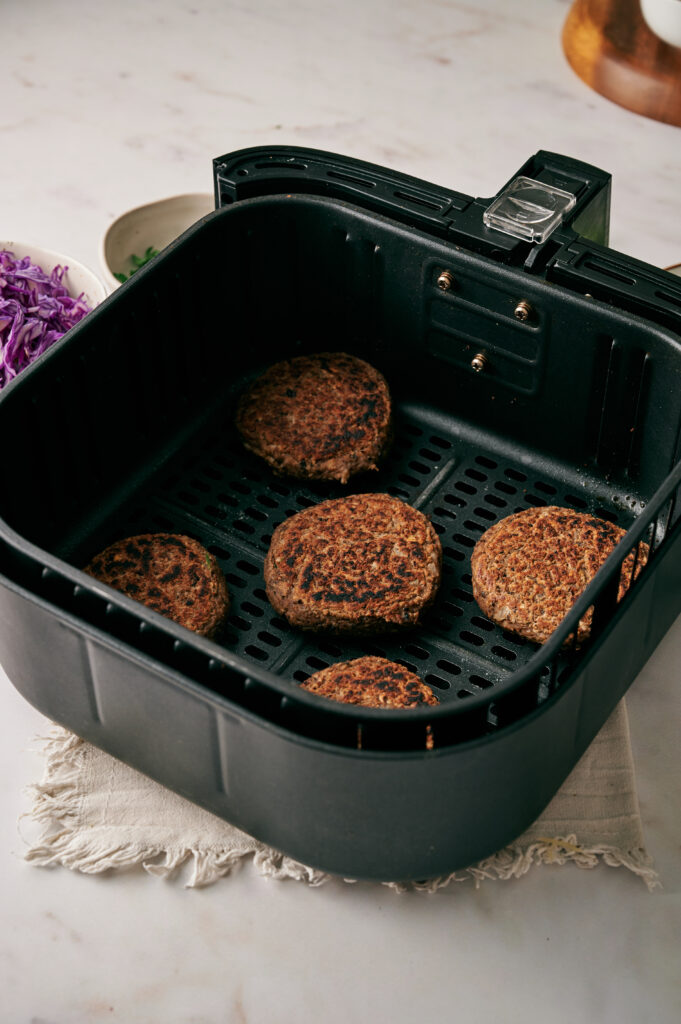 Five Air Fryer Veggie Burger Sliders are arranged inside an air fryer basket, set on a beige cloth atop a light-colored surface.