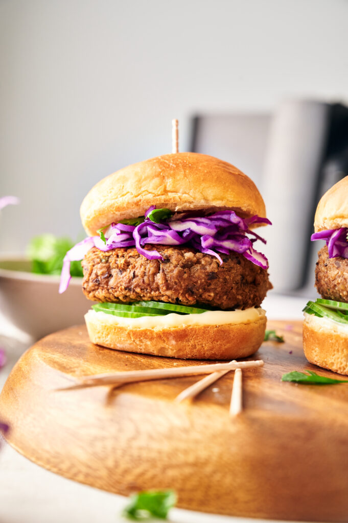An Air Fryer Veggie Burger Slider with a breaded patty, sliced cucumbers, lettuce, mayonnaise, and shredded purple cabbage on a wooden board.