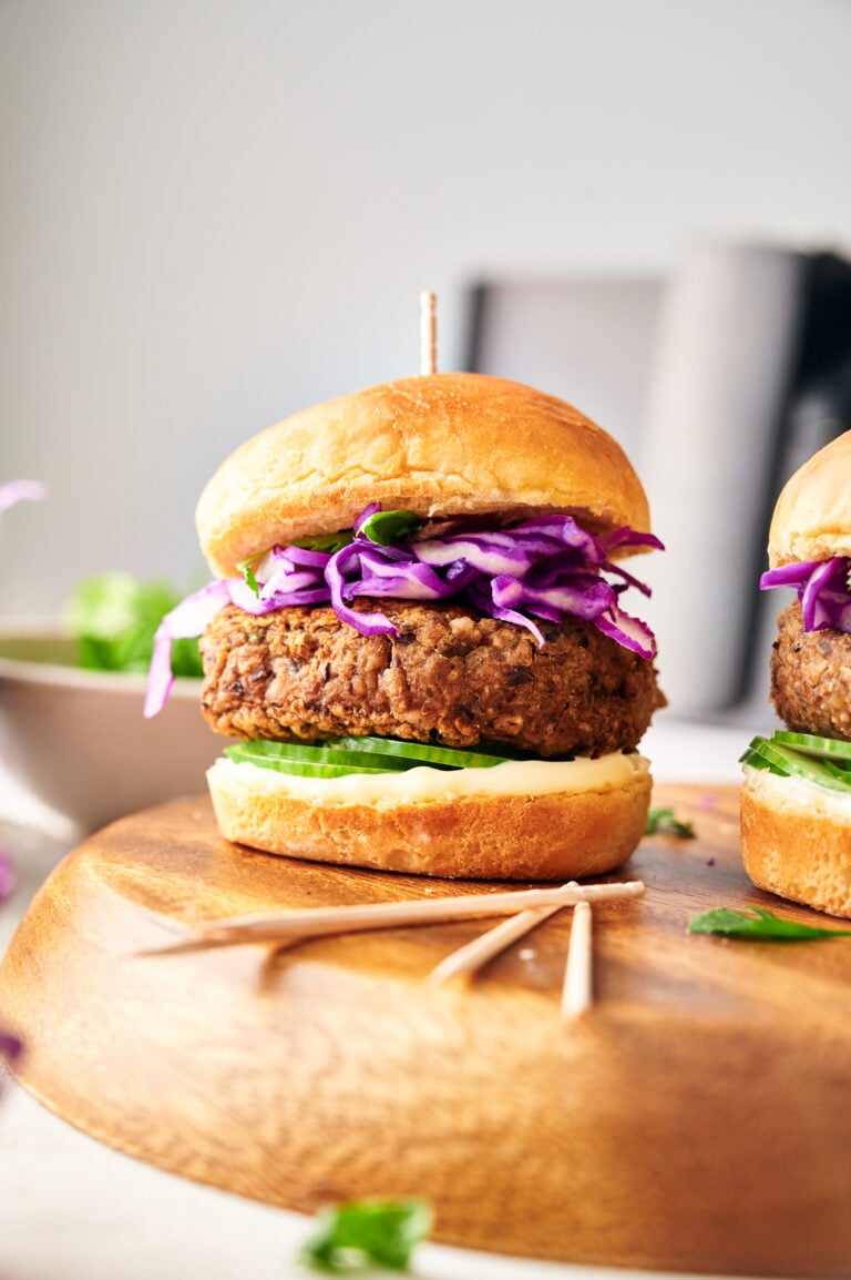 An Air Fryer Veggie Burger Slider with a breaded patty, sliced cucumbers, lettuce, mayonnaise, and shredded purple cabbage on a wooden board.
