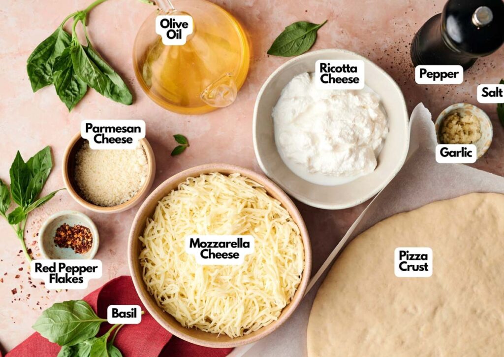 Pizza ingredients arranged on a counter, perfect for making Air Fryer White Pizza, including mozzarella, parmesan, and ricotta cheeses, pizza crust, basil, olive oil, red pepper flakes, garlic, salt, and pepper.