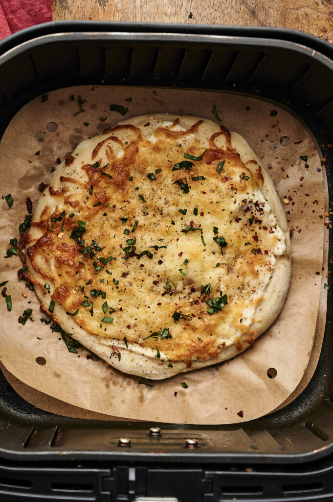 A round flatbread topped with melted cheese and herbs sits on parchment paper inside an air fryer basket, creating a delicious Air Fryer White Pizza ready to be enjoyed.
