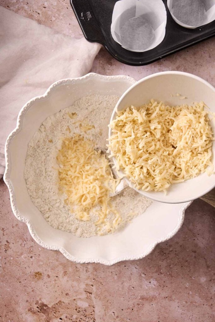 A bowl of grated cheese is being poured into a larger bowl containing flour mixture for Air Fryer Zucchini Muffins, with a muffin tray lined with paper cups in the background.