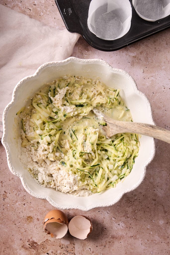 A white bowl with shredded zucchini and batter being mixed for Air Fryer Zucchini Muffins; broken eggshells and a muffin tin are nearby on a stone countertop.