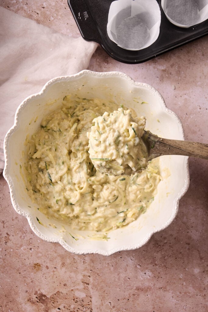 A mixing bowl filled with thick, pale batter for Air Fryer Zucchini Muffins, with shredded zucchini visible and a wooden spoon resting inside. A muffin tray lined with paper cups is partially visible beside it.