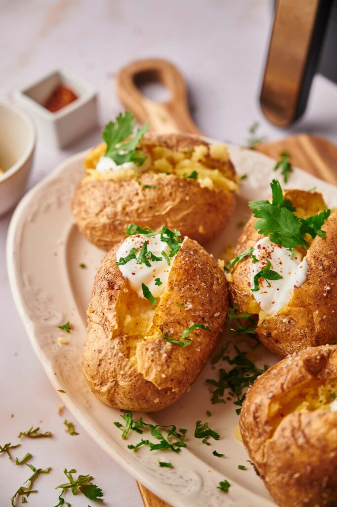 Three air fryer baked potatoes on a plate, topped with sour cream and garnished with chopped parsley and pepper, next to a small container of seasoning.