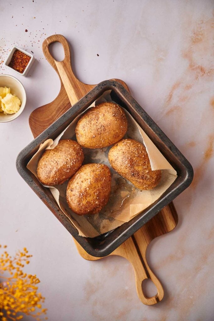A square baking tray with four baked potatoes on parchment paper, placed on a wooden board. A bowl of butter and spices are nearby on a light-colored surface.