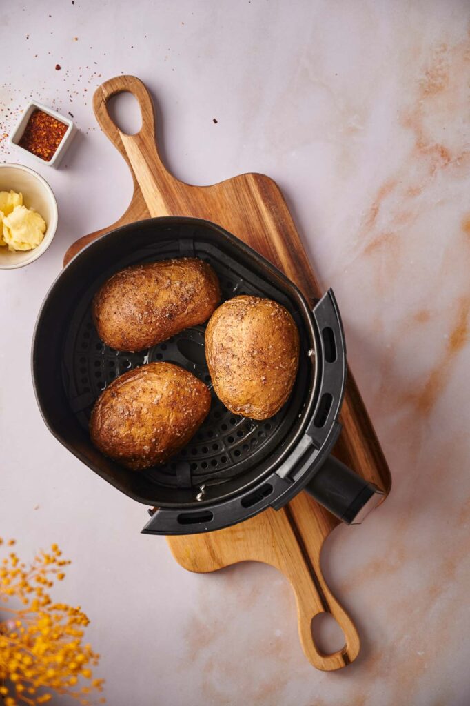Three seasoned Air Fryer Baked Potatoes rest in a basket on a wooden cutting board, accompanied by a bowl of butter and a small dish of spices nearby.