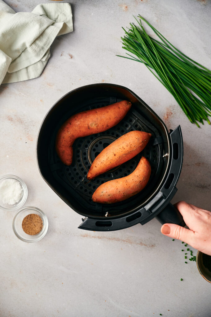 Three whole air fryer sweet potatoes sit in a basket, with a hand holding the handle and ingredients like salt, pepper, and chives scattered on a light countertop.