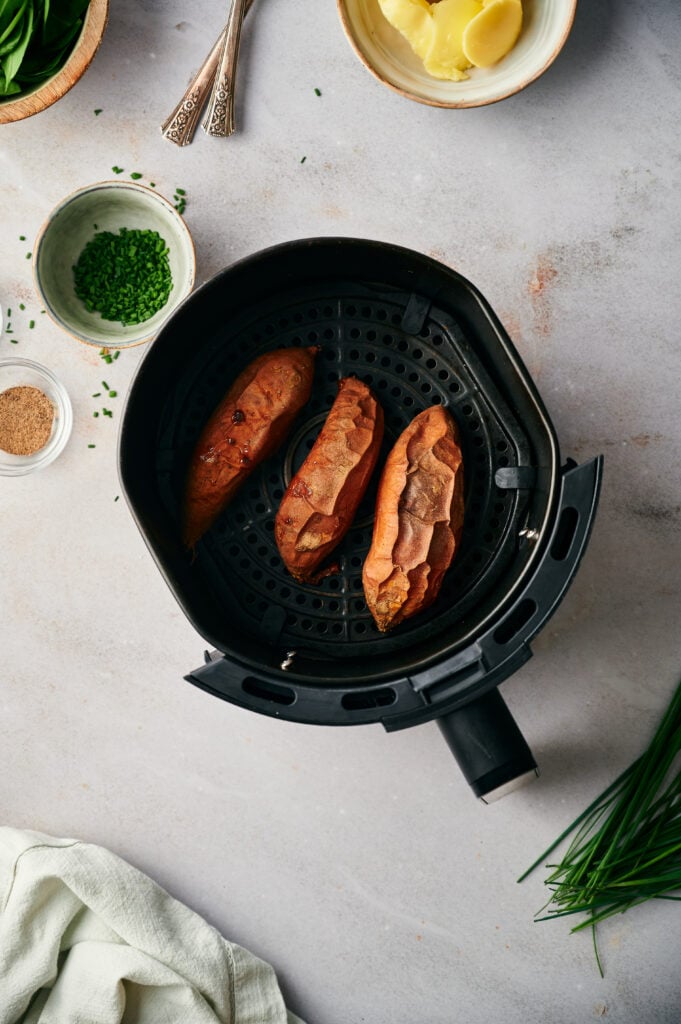 Three Air Fryer Baked Sweet Potatoes rest in a basket, surrounded by bowls of chives, butter, and spices on a light countertop.
