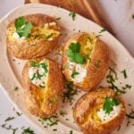 Four baked potatoes on an oval plate, each Baked Potato topped with sour cream, chopped herbs, and a sprig of cilantro, placed on a wooden board.