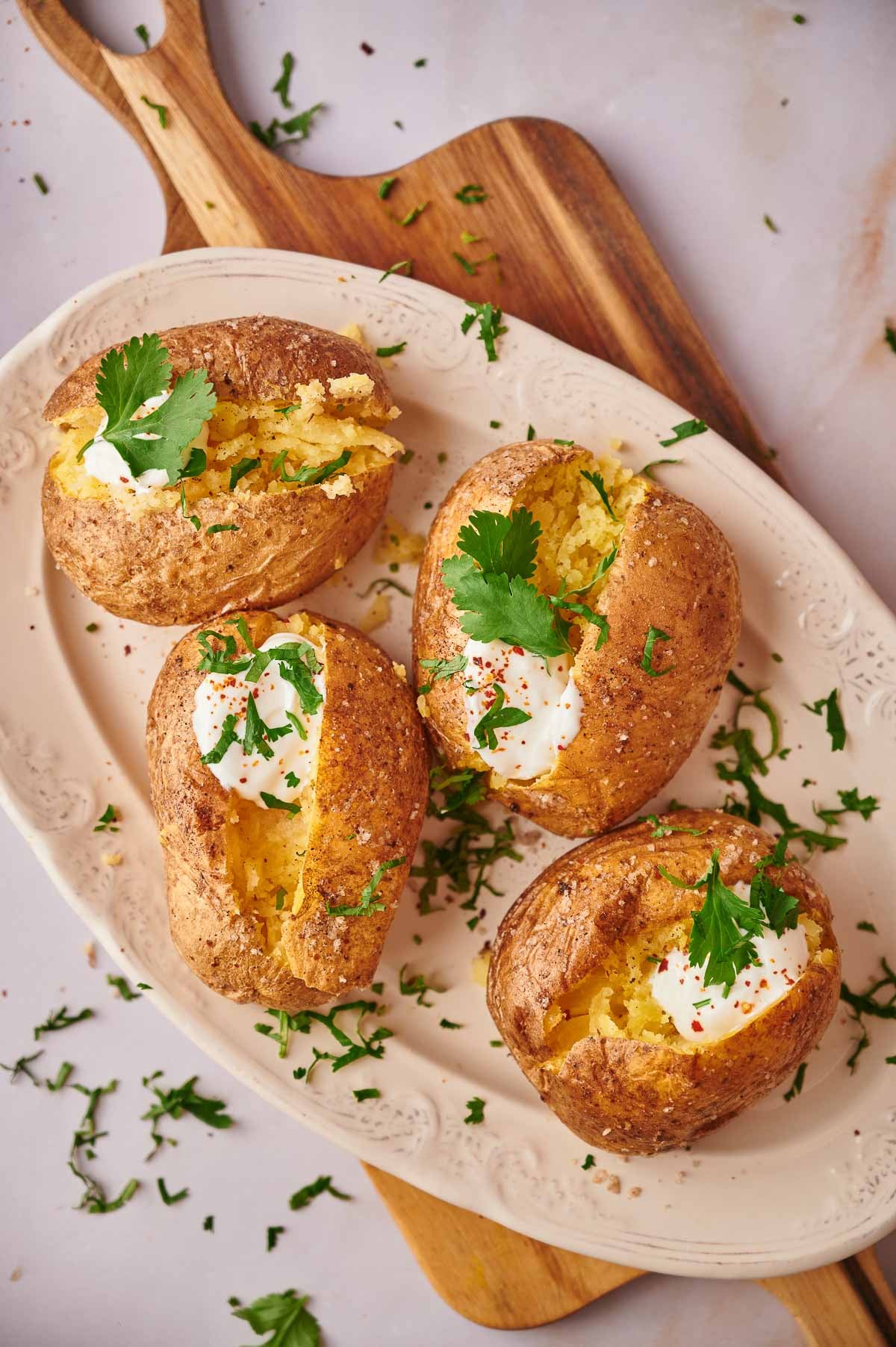 Four baked potatoes on an oval plate, each Baked Potato topped with sour cream, chopped herbs, and a sprig of cilantro, placed on a wooden board.