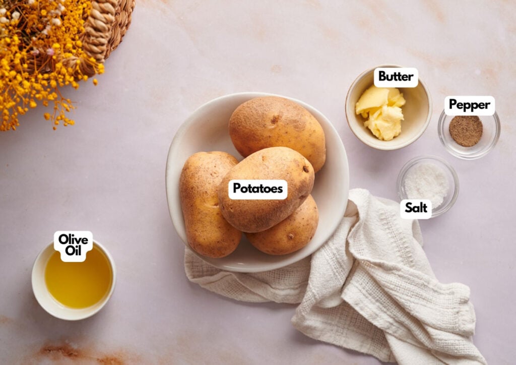A bowl of potatoes, perfect for making air fryer baked potato, is surrounded by small bowls of butter, salt, pepper, and olive oil on a light countertop with a white cloth and a floral arrangement.