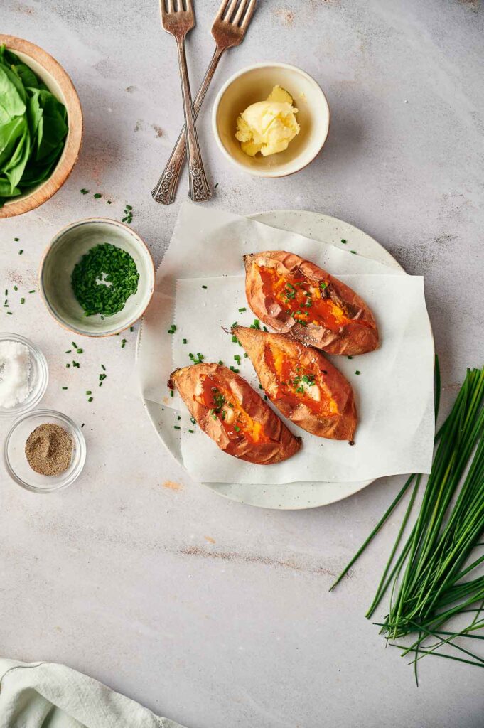 Three air fryer sweet potatoes, baked to perfection and topped with herbs, are on a parchment-lined plate, surrounded by small bowls of butter, salt, pepper, herbs, fresh chives, and two forks.