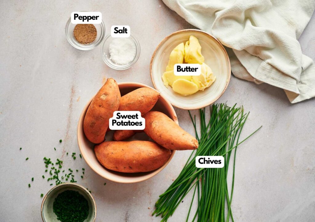 A bowl of Air Fryer Baked Sweet Potato, surrounded by butter, salt, pepper, and fresh chives on a light surface with a cloth nearby.