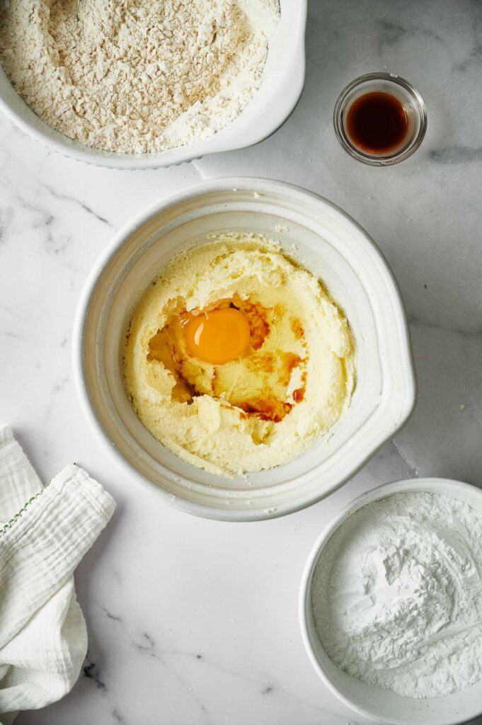 A mixing bowl with creamed butter and sugar, an egg, and vanilla—just the start of delicious Christmas sugar cookies—surrounded by bowls of flour and powdered sugar on a marble countertop.