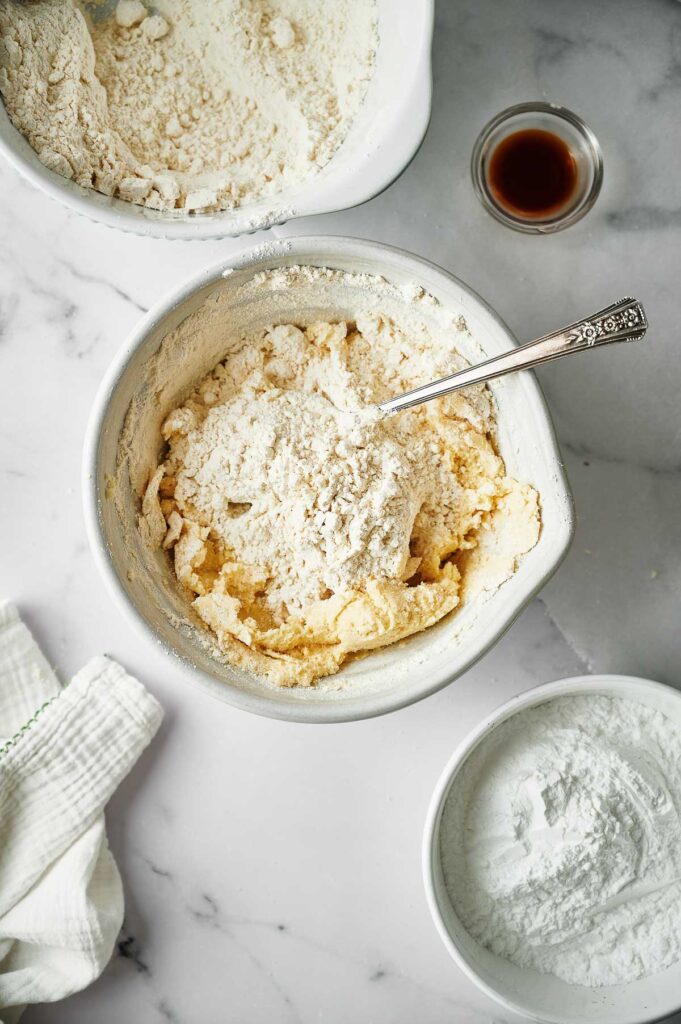 A mixing bowl with flour and batter, a spoon, a bowl of flour, a bowl of powdered sugar, a small cup of vanilla, and a cloth on a marble surface—everything you need for baking classic Christmas sugar cookies.